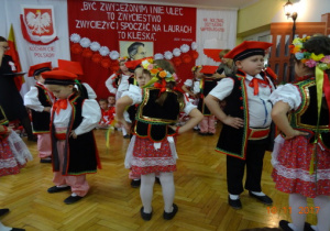 Na tle biało czerwonej dekoracji tańczą w parach dzieci w strojach krakowskich.